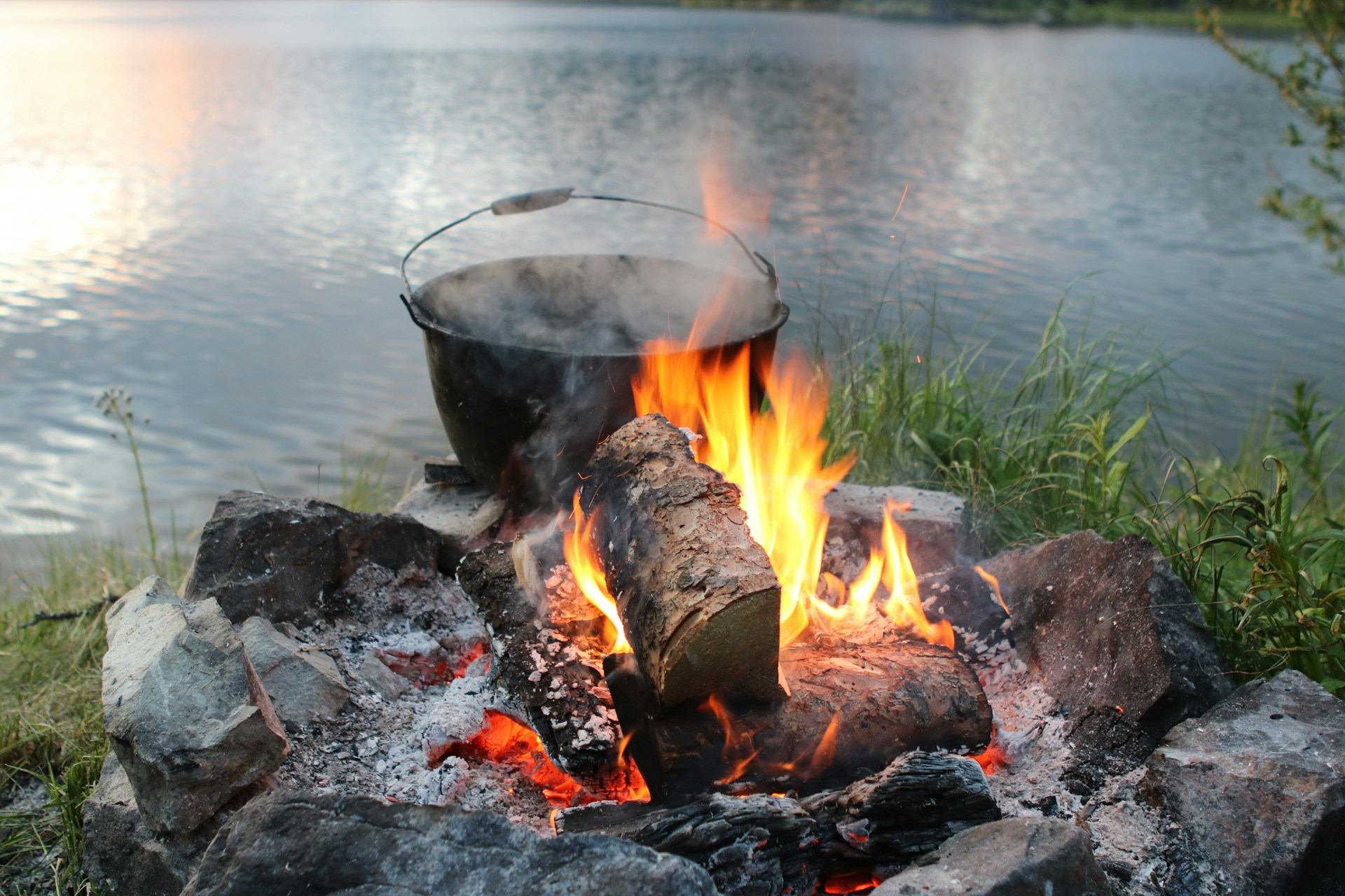 Campfire built with river stones burns beside a calm lake, a blackened cooking pot steaming above the flames in soft evening light.