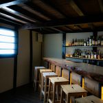 Dimly lit wooden bar with empty stools and shelves of liquor bottles, evoking a quiet, rustic tavern atmosphere.