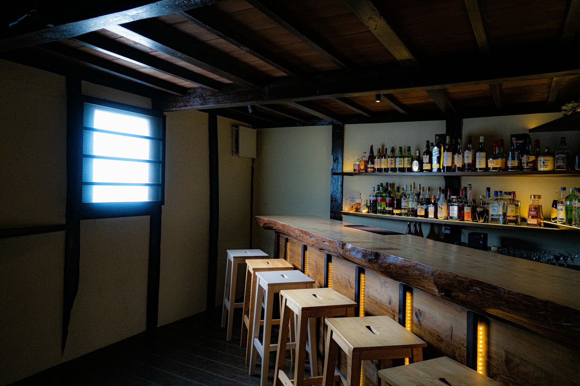 Dimly lit wooden bar with empty stools and shelves of liquor bottles, evoking a quiet, rustic tavern atmosphere.