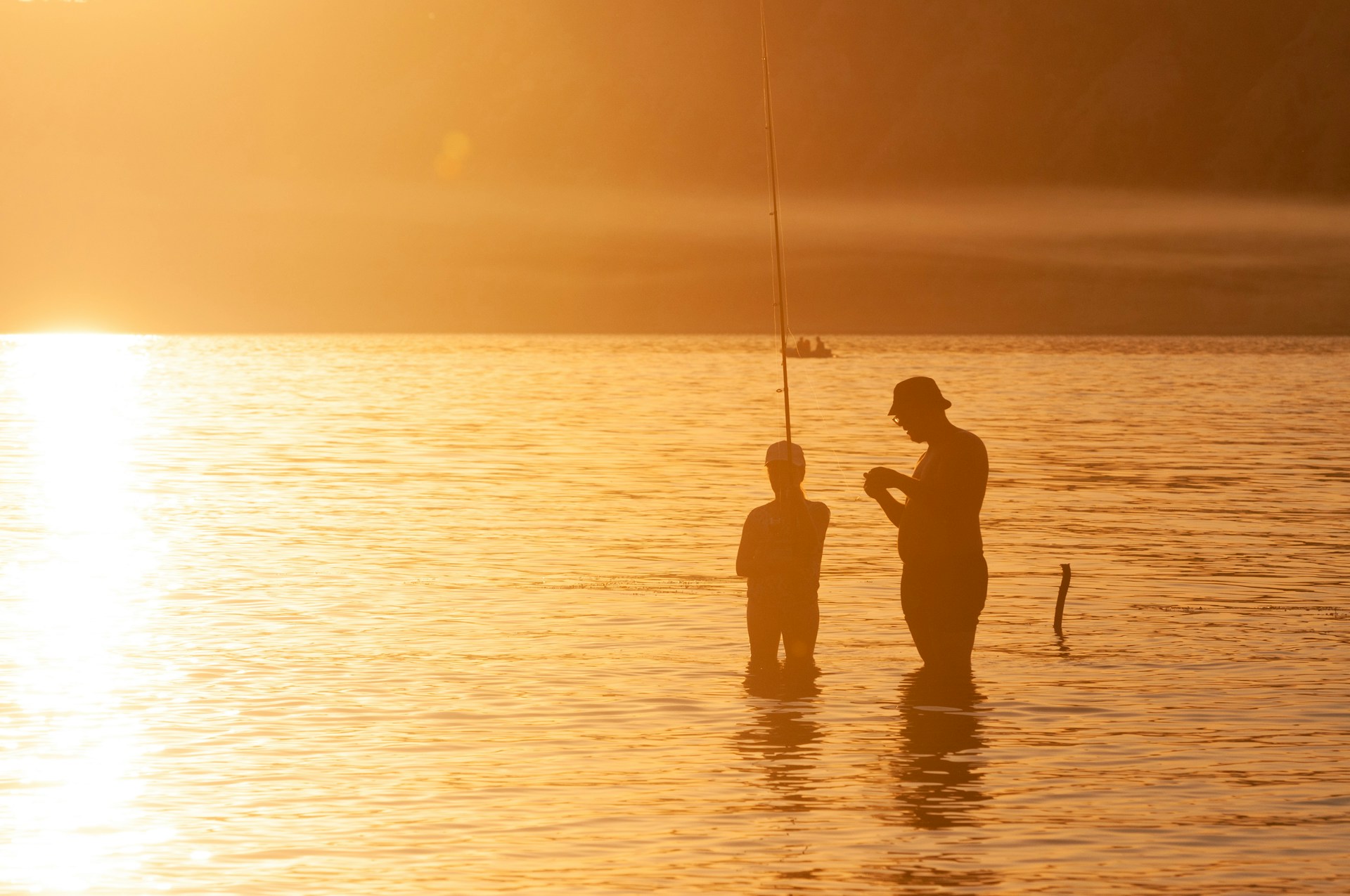 Silhouette of an adult and child standing in shallow water at sunset, fishing together in golden light.