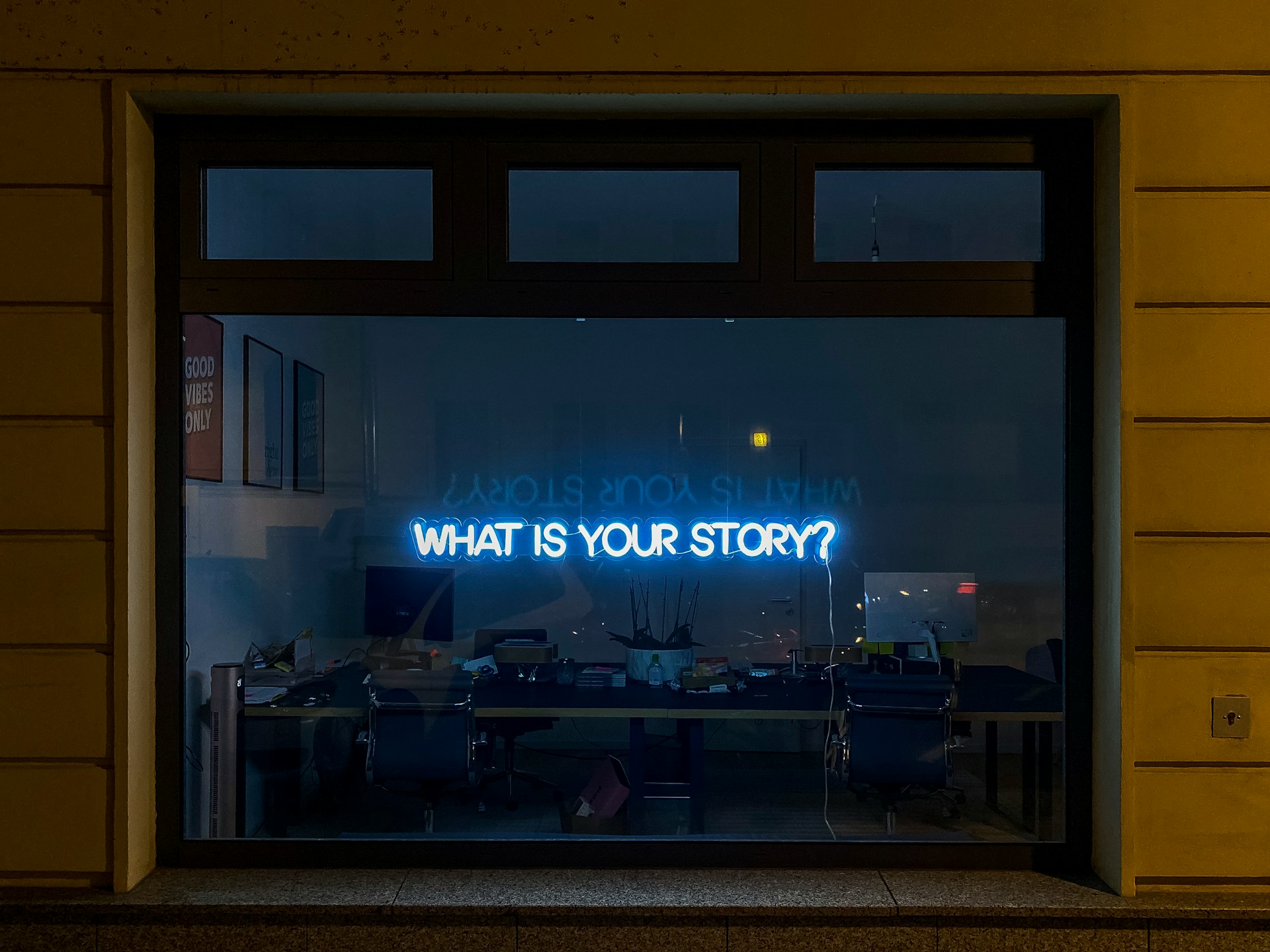 A vibrant blue neon sign asking 'WHAT IS YOUR STORY?' glows suspended in a large office window. The interior is dimly lit, revealing a workspace in productive disarray: dual monitors, stacked books, and a potted plant are visible in the background.